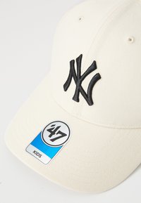 Krém színű baseball sapka fekete hímzett 'NY' logóval, sima anyagból, ívelt karimával és egy kör alakú címkével, amelyen a '47 KIDS' felirat található.