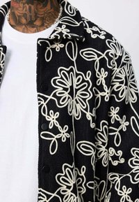 Veste noire à boutons avec broderie florale abstraite blanche portée sur une chemise blanche unie, tatouage sur le cou partiellement visible.