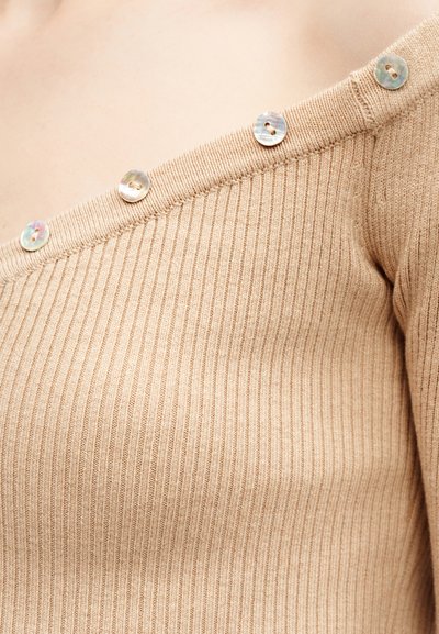 Μπεζ ριγωτό πουλόβερ με ανοιχτούς ώμους και πέντε ιριδίζοντες κουμπιά κατά μήκος της γραμμής του λαιμού, σε ανοιχτό τόνο δέρματος.