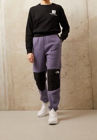 Svart sweatshirt med logotyp, matchat med lila fleecbyxor med svarta knäskydd. Vita sneakers fulländar outfiten.