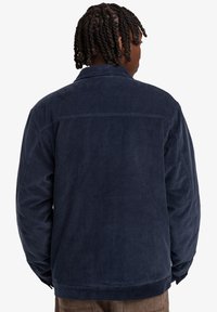 Navy corduroy jacket with a collared neckline and long sleeves. Features textured fabric and a straight hemline at the back.