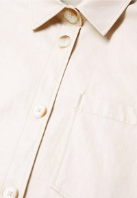 Jasno kremowa koszula z kołnierzykiem, z pionową listwą guzikową i kieszenią na piersi, z czterema okrągłymi guzikami i widocznymi szwami.