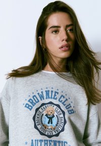 Mujer joven con cabello largo castaño que lleva un sudadera gris con una ilustración de un perro y el texto "Brownie Club Authentic".