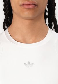 Balta sporta krekls ar apaļu kakla izgriezumu, centrā ir mazs pelēks izšūts Adidas logo. Gluda tekstūra un pieguļošs dizains.