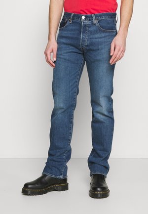 Personne portant un jean bleu droit, des bottes en cuir noires et une chemise rouge, debout devant un fond clair uni.
