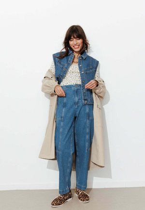 Femme souriante, portant un gilet en jean et un pantalon, un haut à motifs, un trench beige et des chaussures à imprimé léopard, debout contre un mur blanc.