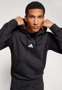 Črna kapuca iz mešanice bombaža, ki ima sprednji žep in bel Adidasov logotip na prsih. Gladka tekstura.