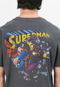 Camiseta de algodón gris con un gráfico colorido de Superman y un villano, con el texto en negrita "SUPERMAN" arriba y detalles de fondo agrietados.