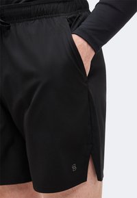 Μαύρα αθλητικά σορτς που φοράει ένα άτομο με το χέρι στην δεξιά τσέπη, ελαστική μέση, μικρό λογότυπο κοντά στο τελείωμα και πλευρική σχισμή στο δεξί πόδι.