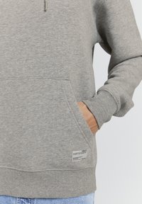 Sudadera gris con una textura suave, cuenta con un bolsillo delantero tipo canguro, capucha con cordón y una pequeña etiqueta en el dobladillo. Diseño simple, corte casual.