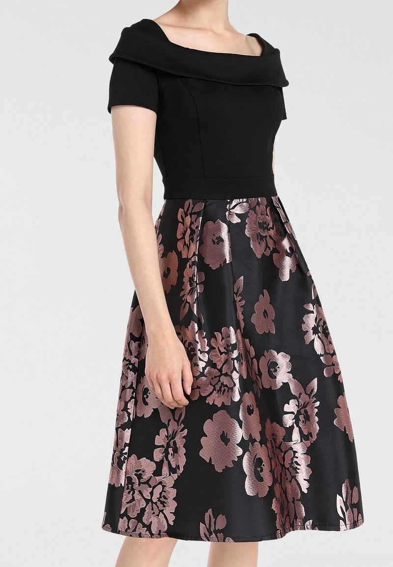 Femme portant une robe noire avec un corsage ajusté à épaules dénudées et une jupe mi-longue ornée de grands motifs floraux roses.