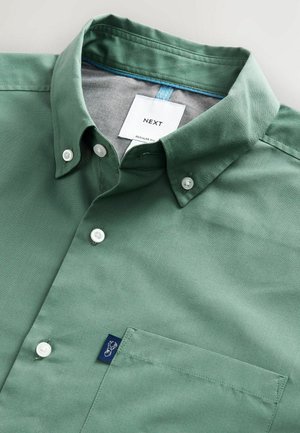 Chemise boutonnée en coton vert avec un col structuré, des boutons blancs et une poche sur le côté gauche de la poitrine présentant une étiquette de logo bleue.
