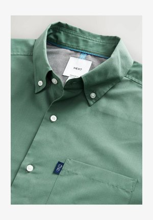 Žalias medvilnės marškiniai su struktūrizuota apykakle, baltais mygtukais ir kairėje krūtinės pusėje esantimi kišene su mėlynu logotipo ženkleliu.