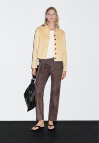 Beige wool jacket with black buttons, white top, brown suede pants, and a large black tote bag. Flip-flops complete the outfit.