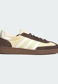 Χαμηλού προφίλ μπεζ και καφέ αθλητικό sneaker Adidas με λευκές ρίγες, υφασμένο πέλμα και σουέντ διακοσμήσεις σε ανοιχτό φόντο.
