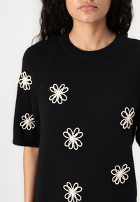 Maglione nero a maniche corte con disegni floreali ricamati bianchi su tutto il capo. Il collo è a coste per un tocco di texture.