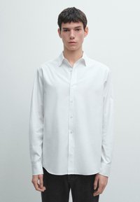 Camicia bianca a maniche lunghe in tessuto liscio, con colletto con bottoni e taglio dritto, abbinata a pantaloni scuri.