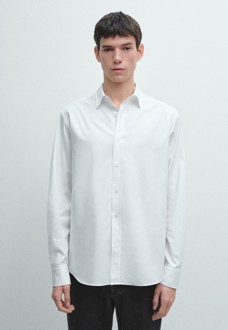 Camicia bianca a maniche lunghe in tessuto liscio, con colletto con bottoni e taglio dritto, abbinata a pantaloni scuri.