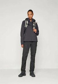 Must värvi välistingimustes jope, millel on kapuuts, sobib kokku mustade pükstega ja kerge seljakotiga, millel on valged rihmad, seistes ühetoonilisel taustal.