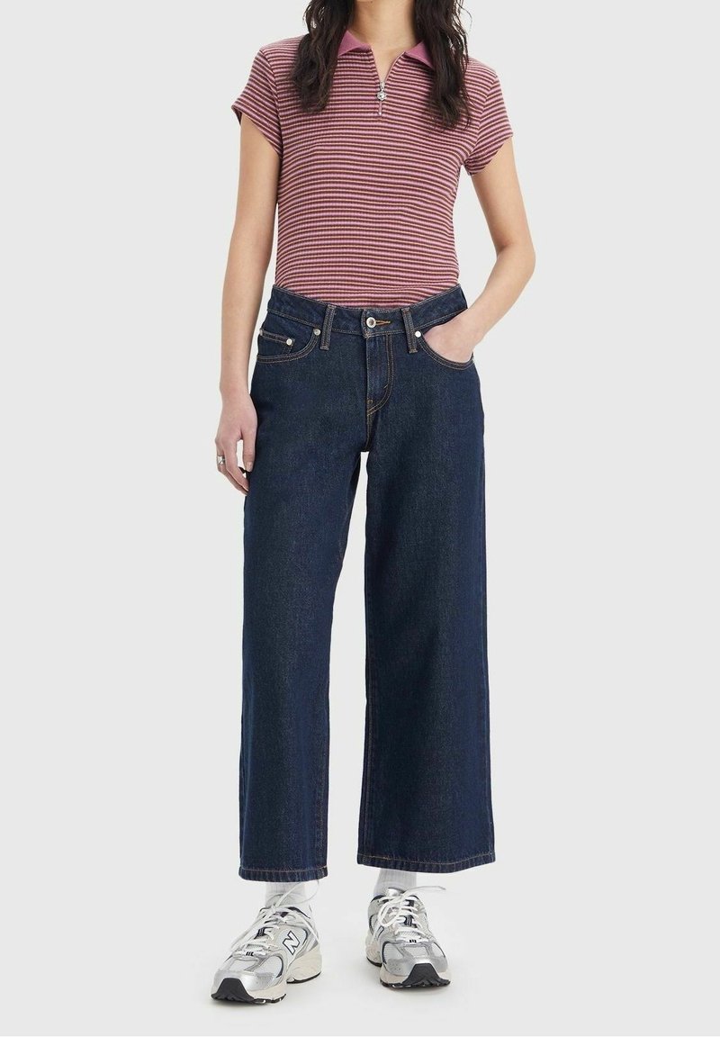 Polo rayé rouge et rose avec une fermeture éclair, associé à un jean en denim bleu foncé à jambes larges et des baskets de sport argentées.
