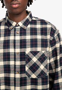 Camisa de franela a cuadros en crema, azul marino y burdeos. Presenta un collar, cierre de botones y un bolsillo en el pecho con un patrón diagonal.