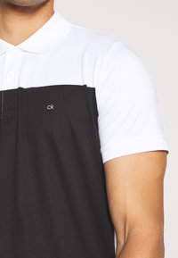Homme portant un polo noir et blanc à manches courtes avec le logo "ck" sur la poitrine.