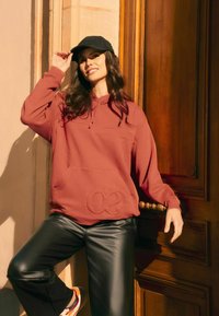 Weinrote Kapuzenjacke mit erhabenem "30"-Design, Zugkordel am Hals und Fronttasche, kombiniert mit schwarzen Lederhosen und einer schwarzen Mütze.