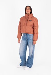 Brązowa krótka kurtka puchowa z wysokim kołnierzem, teksturowanym wzorem i naszywką z logo, w połączeniu z luźnymi niebieskimi dżinsami i białymi trampkami.