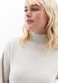 Hellbeige gerippte Strick-Turtleneck-Oberteil mit strukturiertem Kragen und lockerem Schnitt, das eine weiche Textur und subtile vertikale Linien aufweist.