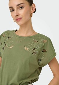 T-shirt vert olive à manches courtes en coton, avec des découpes florales à l'avant et une coupe décontractée.