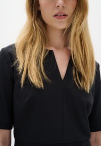 Juoda suknelė su trumpomis rankovėmis ir V formos iškirpte, pagaminta iš lygaus audinio, su subtiliomis siūlėmis ir be matomų raštų ar metalinių detalių.