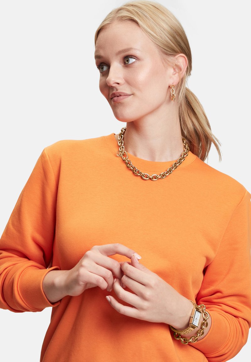 Orange sweatshirt med lange ærmer, featuring en rund halsudskæring. Guld kæde halskæde og armbånd tilføjer metalliske accenter. Glat tekstur.