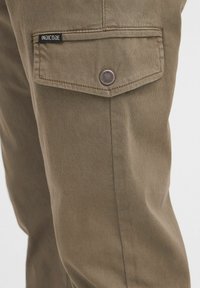 Khaki spodnie cargo z boczną kieszenią z klapką i zapięciem na zatrzask; zawierają metkę "Indicode" na kieszeni.