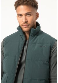 Dunkelgrüne gepolsterte Weste mit hohem Kragen, Reißverschluss und Logodetails. Über einem grauen Langarmshirt getragen. Glatte Stofftextur.
