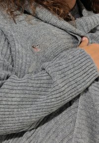 Šedý pletený svetr s žebrovanou texturou, s mírným roztržením, vysokým límcem a překrývajícími se látkovými detaily.