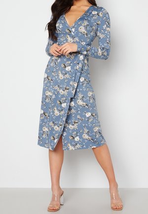 Sukienka kopertowa w jasnoniebieskim kolorze, z długimi rękawami, głębokim dekoltem w serek i bocznym rozcięciem. Tkanina ma miękką fakturę z żywym, kwiatowym wzorem.