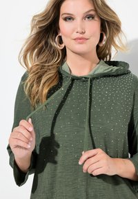 Sweat à capuche vert en tissu texturé, orné de clous en argent à l'avant, avec un cordon de serrage et des manches amples.