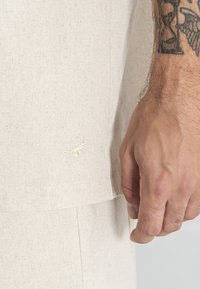 Camisa de lino beige con detalle de bordado de escorpión. La tela tiene una textura sutil, con un corte recto y mangas cortas.