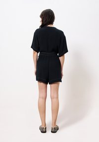 Schwarzer Kurzarm-Overall aus strukturiertem Stoff mit tailliertem Gürtel, kontrastierenden Nähten und Shorts über dem Knie.