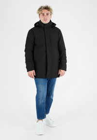 Giacca imbottita nera con collo alto e cappuccio, dotata di chiusura lampo frontale, tasche laterali e dettagli arancioni a contrasto. Indossata su jeans blu e sneakers bianche.