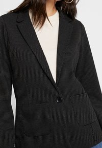 Svart blazer med vita prickar, kavajslag, enfärgad knäppning och sidofickor; tyget verkar slätt med en strukturerad passform.