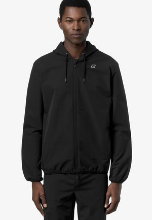 Giovane uomo che indossa una giacca nera con cappuccio e zip con logo sul petto e pantaloni neri abbinati, in piedi davanti a uno sfondo bianco.