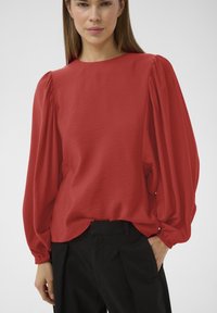 Femme portant un chemisier rouge à manches longues bouffantes et un pantalon noir taille haute, debout avec une main dans la poche.