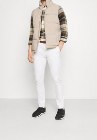 Gilet imbottito beige con colletto alto, camicia a quadri nei colori verde, marrone e bianco, pantaloni bianchi slim e scarpe da ginnastica nere.
