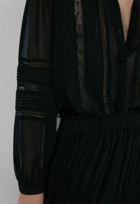 Blouse noire avec manches transparentes, détails en dentelle, rayures verticales et taille froncée. Tissu texturé avec fermeture boutonnée à l'avant.