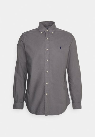 Camisa de botones en tela gris oscuro con mangas largas, cuello puntiagudo y pequeño logo en el pecho. Presenta botones de color claro.