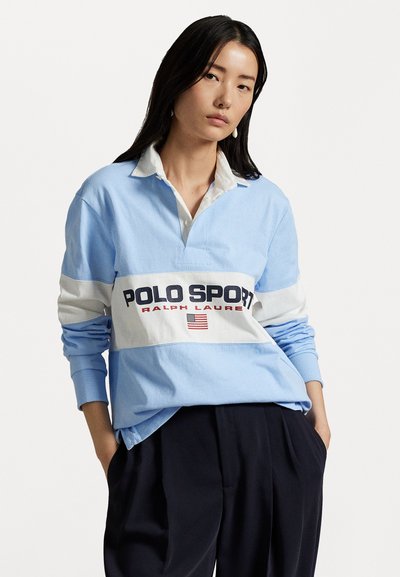 Gaiši zils garām piedurknēm polo krekls ar baltu paneli, uz kura ir melnā un sarkanā krāsā uzraksti "POLO SPORT" un "RALPH LAUREN", papildināts ar mazu Amerikas karogu.