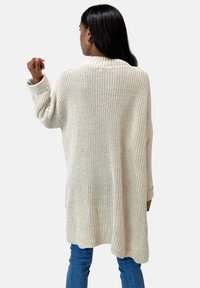 Vrouw met lang zwart haar, gekleed in een crèmekleurige chunky gebreide cardigan en blauwe jeans, die met de rug naar ons gekeerd staat en haar linkerarm licht omhoog steekt.
