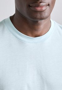 Šviesiai mėlynas medvilninis marškinėlis su apvaliu kaklo iškirpimu, lygiu paviršiumi ir be matomų raštų ar akcentų. Artimas kaklo ir viršutinio audinio vaizdas.