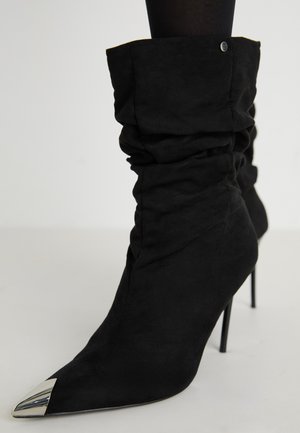 Sorte slouchy ankelstøvler med spiss tå og metallisk sølvdetalj. Laget av mykt semsket skinn, med en høy stilettahæl.